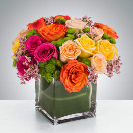 Compact arrangement of orange, yellow, pink and peach roses with green accents in a clear glass cube vase.