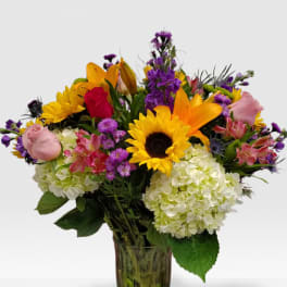 Tall mixed bouquet with sunflowers, roses, lilies, hydrangeas and purple flowers in a clear glass vase