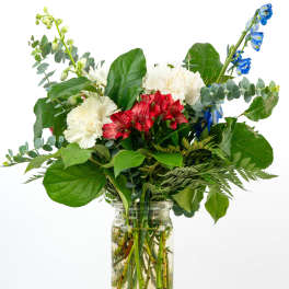 Red, white, and blue mixed flower arrangement with lush foliage in a clear glass jar vase