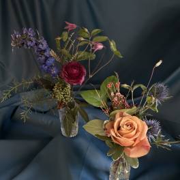 Two small bud vase arrangements with a peach rose, purple blooms, and berries on a dark teal fabric backdrop