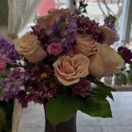 Peach roses with purple and pink blooms arranged in a ribbed purple glass vase