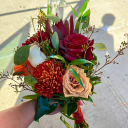 Handheld bouquet of red, peach, and burgundy flowers with greenery