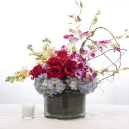Low arrangement of red roses, purple orchids, and blue hydrangeas in a glass vase with a small white candle.