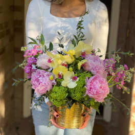 Lush arrangement of pink peonies, yellow orchids and green hydrangeas in a gold pot held by a person in white.