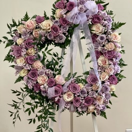 Heart-shaped wreath of pink and lavender roses on a white stand with ribbon