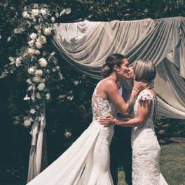 Two brides kissing beneath a floral wedding arch with draped fabric