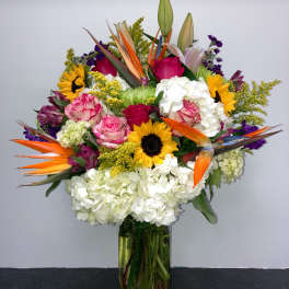 Colorful mixed bouquet in a clear glass vase with sunflowers, roses, lilies, and hydrangeas