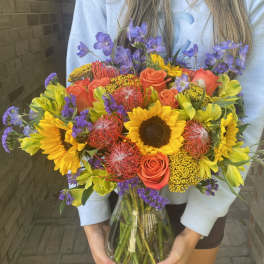 Bright mixed bouquet with sunflowers, orange roses, and purple flowers in a glass vase