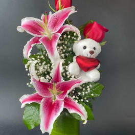 Pink lilies and red roses in a green vase with a teddy bear holding a heart