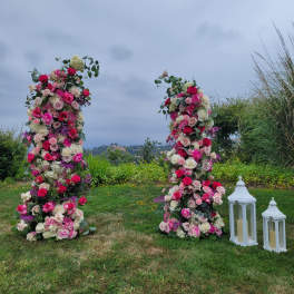 Two tall outdoor floral columns of pink and white roses with white lanterns on the grass