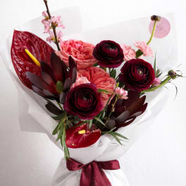 Hand-tied bouquet of dark red ranunculus, pink roses, and tropical blooms wrapped in white paper with burgundy ribbon