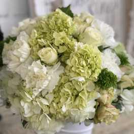 Round arrangement of white and pale green hydrangeas, roses, and carnations in a white vase