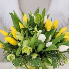 Yellow and white tulip arrangement with green blooms in a round vase with ivy and a yellow ribbon bow.