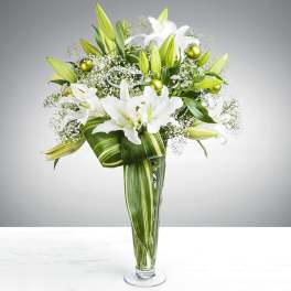 Tall glass vase of white lilies with baby's breath, green ornament balls, and a large variegated leaf bow.