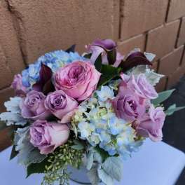 Low vase arrangement of pink and lavender roses, blue hydrangeas, and dark purple calla lilies.