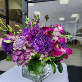 Compact arrangement of purple and pink flowers in a clear glass cube vase.