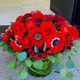 Low arrangement of vivid red flowers with dark centers in a leaf-wrapped glass vase