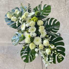 Tall standing spray of white roses and mums with large dark green monstera leaves on an easel