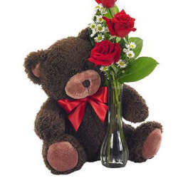 Brown teddy bear with red bow beside a clear vase of red roses and small white daisies.