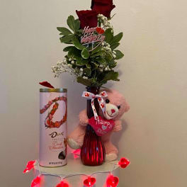 Two red roses in a vase with a teddy bear, chocolate tin, and heart lights