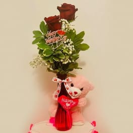 Red roses in a red vase with a pink teddy bear and Valentine sign