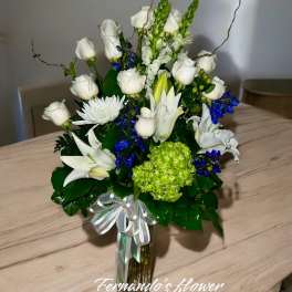 Tall arrangement of white roses and lilies with blue accents in a glass vase with a ribbon bow.