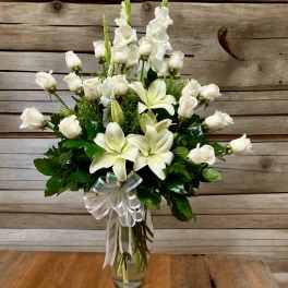 Tall clear vase of white roses, lilies, and snapdragons with a white ribbon bow.