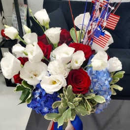 Patriotic red, white, and blue bouquet with roses and hydrangeas in a glass vase with American flags.