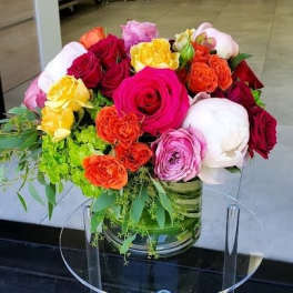Low glass vase of bright roses, peonies, and spray roses in pink, red, orange, and yellow on a clear stand.