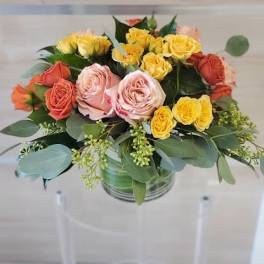 Round arrangement of yellow, peach, and orange roses in a clear glass cylinder vase