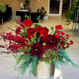Low, lush arrangement of red roses and deep red flowers with evergreens in a gold container on a clear stand