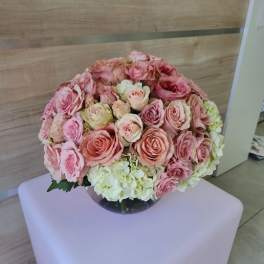 Round arrangement of pink roses and white hydrangeas in a clear glass bowl vase on a pale cube stand