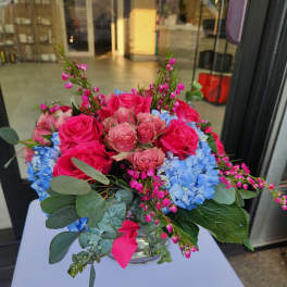 Low glass vase arrangement with hot pink roses and blue hydrangeas accented by small pink blossoms.