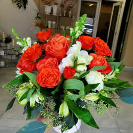 Arrangement of bright orange roses and white flowers with green foliage in a white round vase.