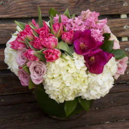 Low round bouquet of white hydrangeas, pink roses, tulips, and purple orchids in a clear glass vase.