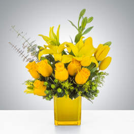 Yellow lilies, roses, and tulips arranged in a yellow glass vase with green berries