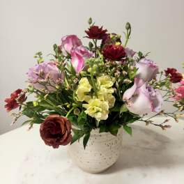 Mixed bouquet of pink, lavender, yellow, and burgundy flowers in a white vase