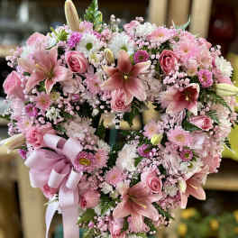 Heart-shaped standing wreath of pink lilies, roses, and mums with a large pink ribbon bow.