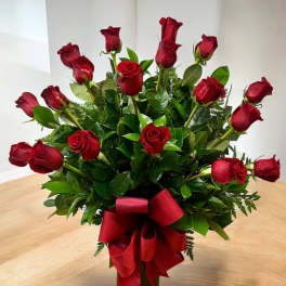 Tall arrangement of long-stem red roses with lush foliage in a dark vase with a red bow
