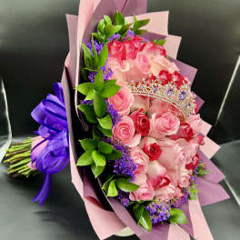 Bouquet of pink and red roses with purple filler flowers, wrapped in pink paper with a small tiara decoration.