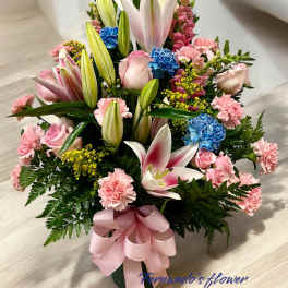 Tall arrangement of pink lilies, roses, carnations, and blue flowers with a pink bow in a green vase