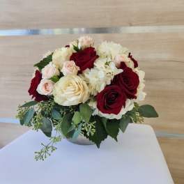 Low round arrangement of red and cream roses, blush spray roses, and white hydrangeas in a small bowl vase.