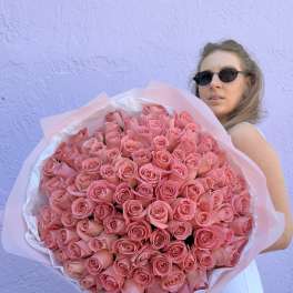 Large bouquet of tightly clustered pink roses wrapped in paper, held by a woman in sunglasses.