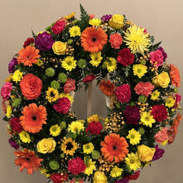 Bright circular wreath of orange gerbera daisies, yellow roses, and mixed flowers on a wooden easel.