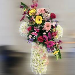 Floral cross of white mums adorned with pink roses, gerberas, lilies and colorful butterfly accents.