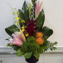 Tropical arrangement with pink lilies, orange pincushion blooms, and yellow orchids in a dark vase