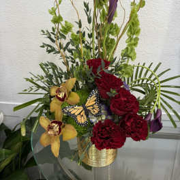 Tall floral arrangement with red roses, purple calla lilies, and yellow orchids in a gold vase