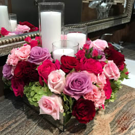 Pink, red, and lavender roses arranged around white pillar candles in glass holders.