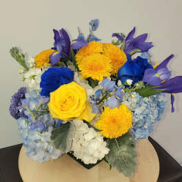 Blue and yellow floral arrangement with roses, hydrangeas, and daisies in a low container