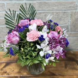 Round arrangement of pink roses, pink carnations, white alstroemeria, and purple blooms in a clear glass vase
